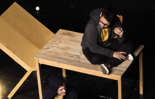 Diapositive pour le spectacle L'envers de nos décors de Clément Dazin et Thomas Scotto. Un homme est assis sur une table en pin avec des boules de jonglages partout autour de lui.