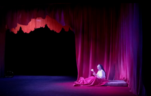 Photo du spectacle de Jeanne Lepers "Le bon fruit mûr" avec le personnage féminin portant un masque, allongée dans son lit, devant un rideau rouge.