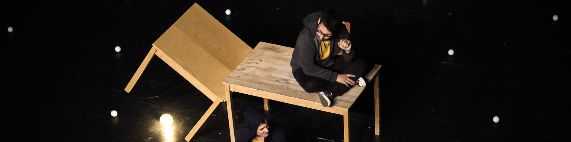 Diapositive pour le spectacle L'envers de nos décors de Clément Dazin et Thomas Scotto. Un homme est assis sur une table en pin avec des boules de jonglages partout autour de lui.