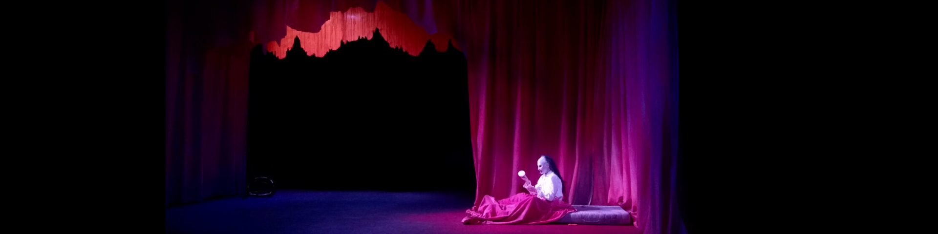 Photo du spectacle de Jeanne Lepers "Le bon fruit mûr" avec le personnage féminin portant un masque et allongée dans un lit devant un rideau rouge.